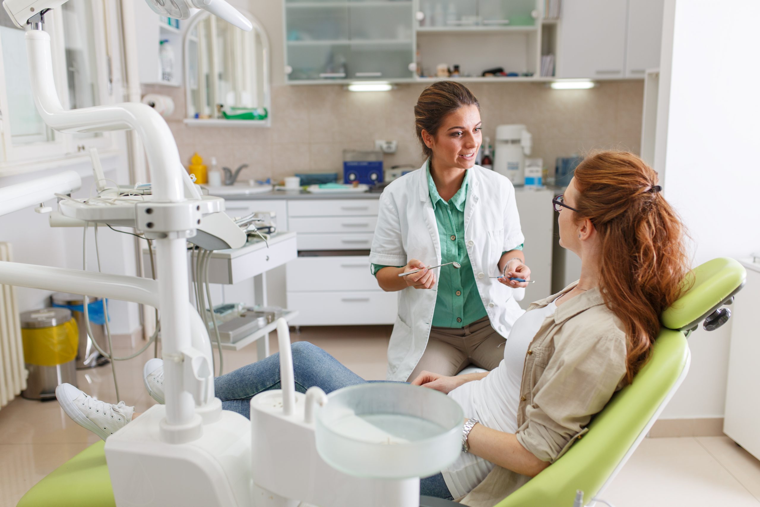  A female dentist holds dental tools and talks to a female patient sitting in an exam chair. 