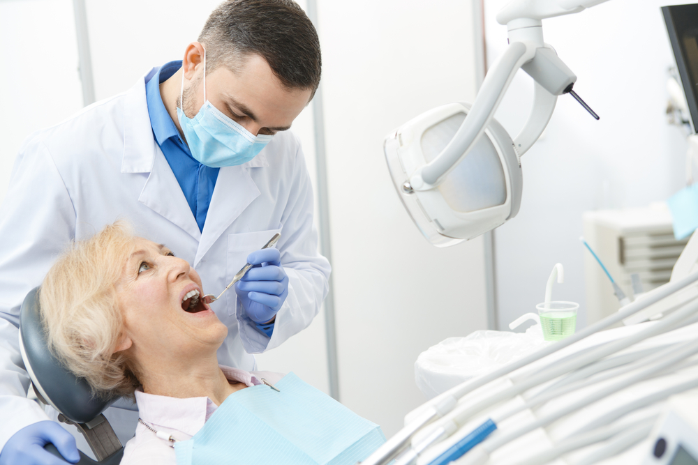 A male dentist uses a dental tool with an older female patient. 