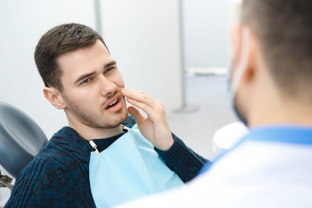 Man reclining in dental chair, wearing dental bib, holds left hand to mouth, indicating to dentist where he feels tooth pain.