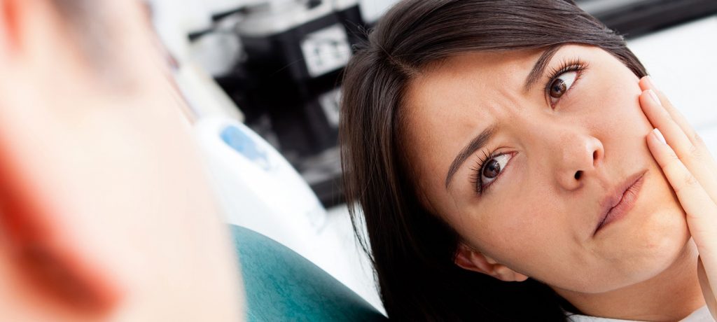 Woman reclining in dental chair looks at dentist with left hand to her cheek, indicating where she feels tooth pain. 
