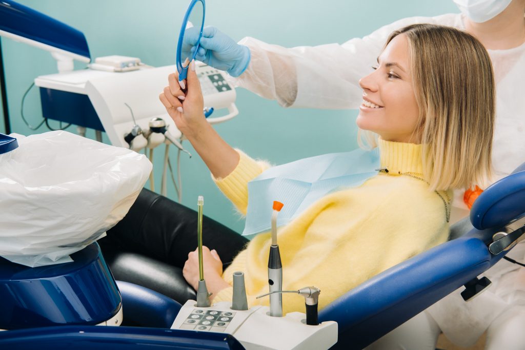 A young woman with blonde short hair admires her teeth whitening results in a handheld mirror held by dentist. 