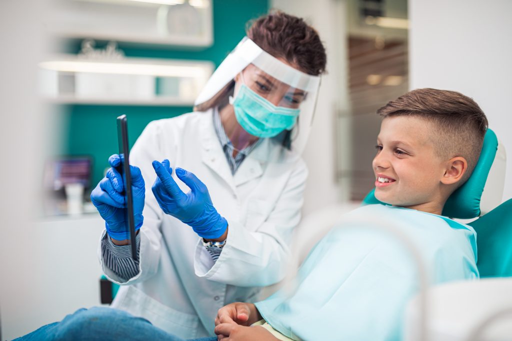 A female dentist in personal protective equipment (PPE) shows dental tool to male boy patient in dental chair.