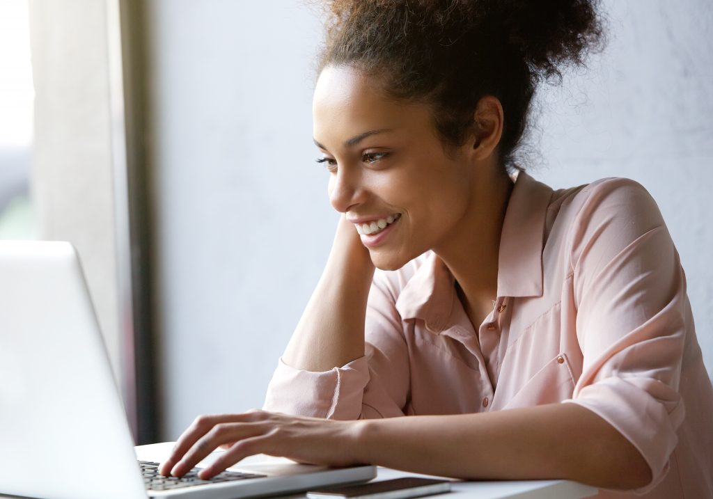 A beautiful black woman in pink shirt with her hair pulled back, searches for how to choose a dentist on her laptop.