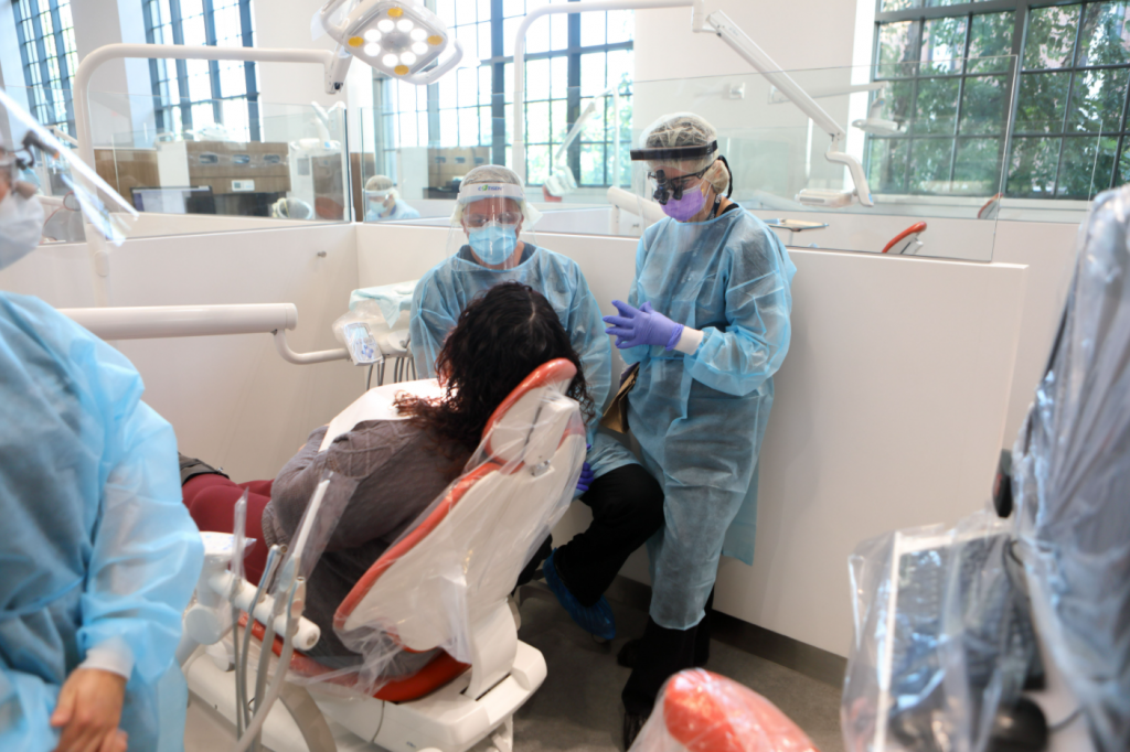 A group of student dentist wearing facemask, face shields and gloves performs talk about a root canal to a female patient.