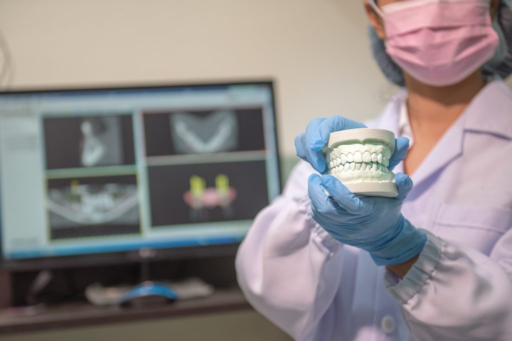 A female prosthodontist in personal protective equipment (PPE) holds model of a mouth with X-rays in background. 