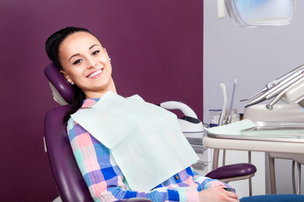  A young woman with dark brown hair and eyes sits comfortably in dental chair, not feeling pain after root canal. 
