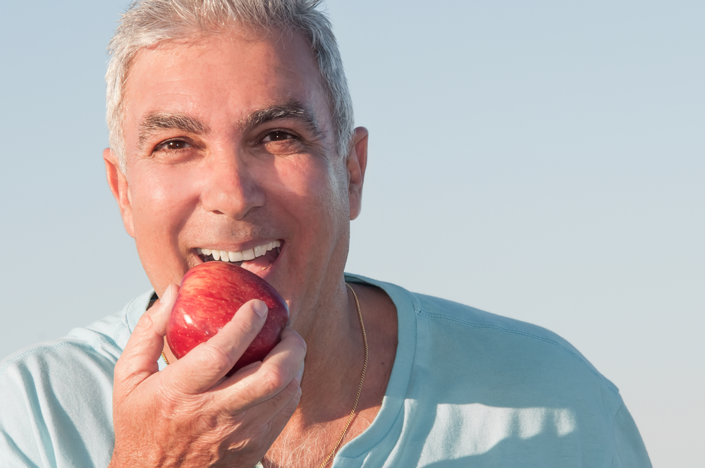  An older man smiles because he can eat an apple now that he’s undergone the dental implant process. 