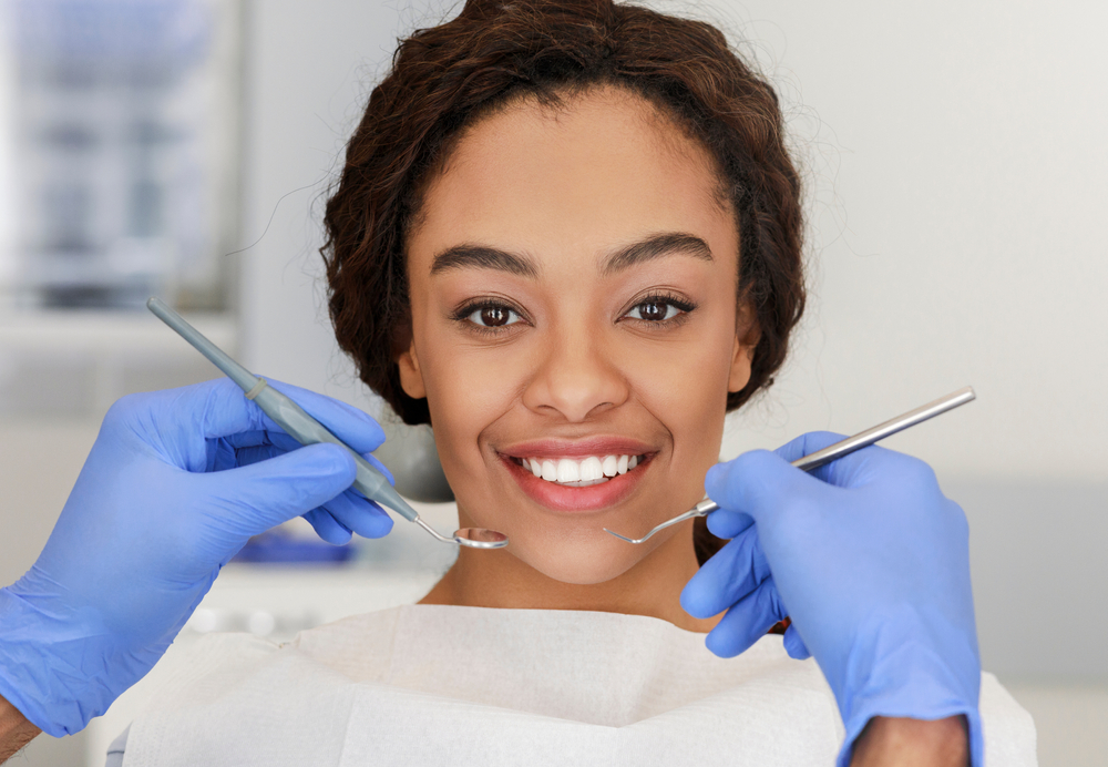 A girl in an exam chair at a dentist’s office smiles while the dentist holds tools. 