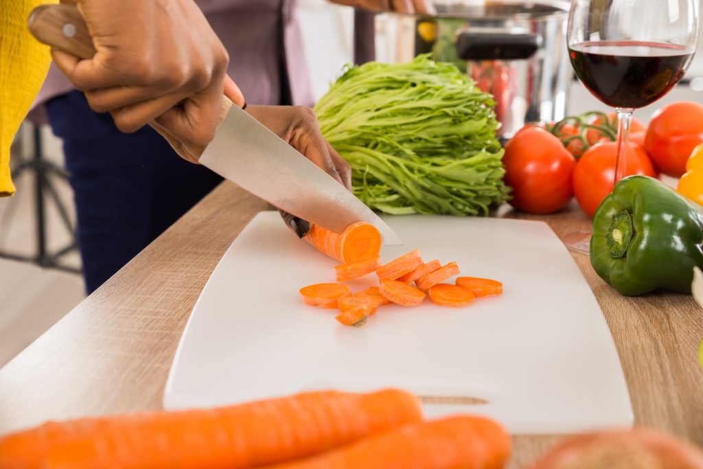 A closeup image of a woman’s hand cutting up carrots for a healthy salad with a glass of wine on the counter. 