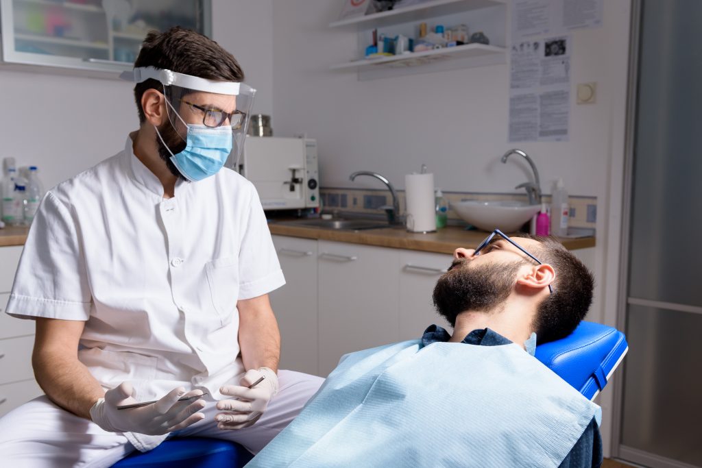A male dentist in personal protective equipment (PPE) provides routine dental care for a male patient with glasses. 
