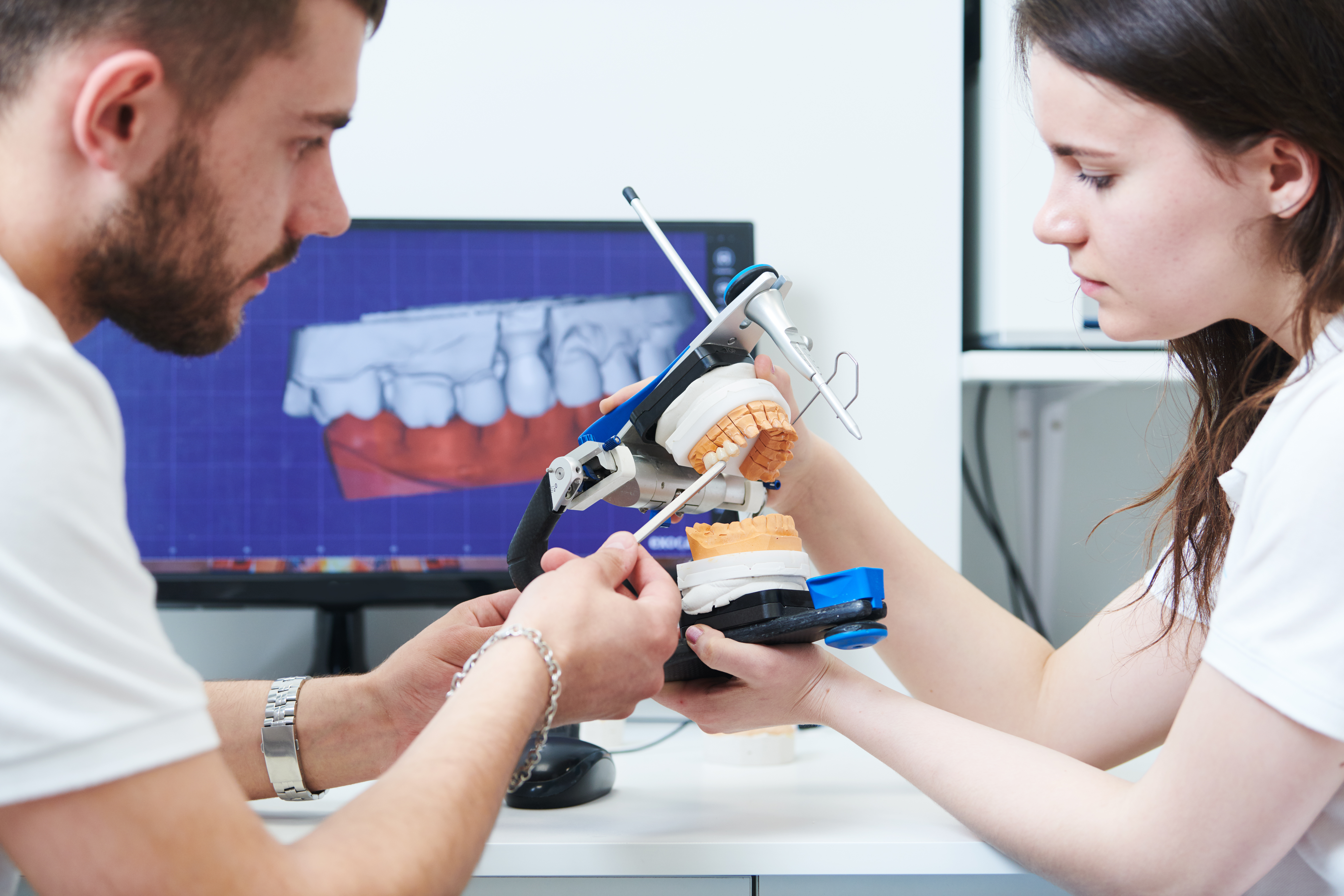 Two dentistry students use tool to examine dental prosthodontics, digital image of teeth on computer monitor in background. 