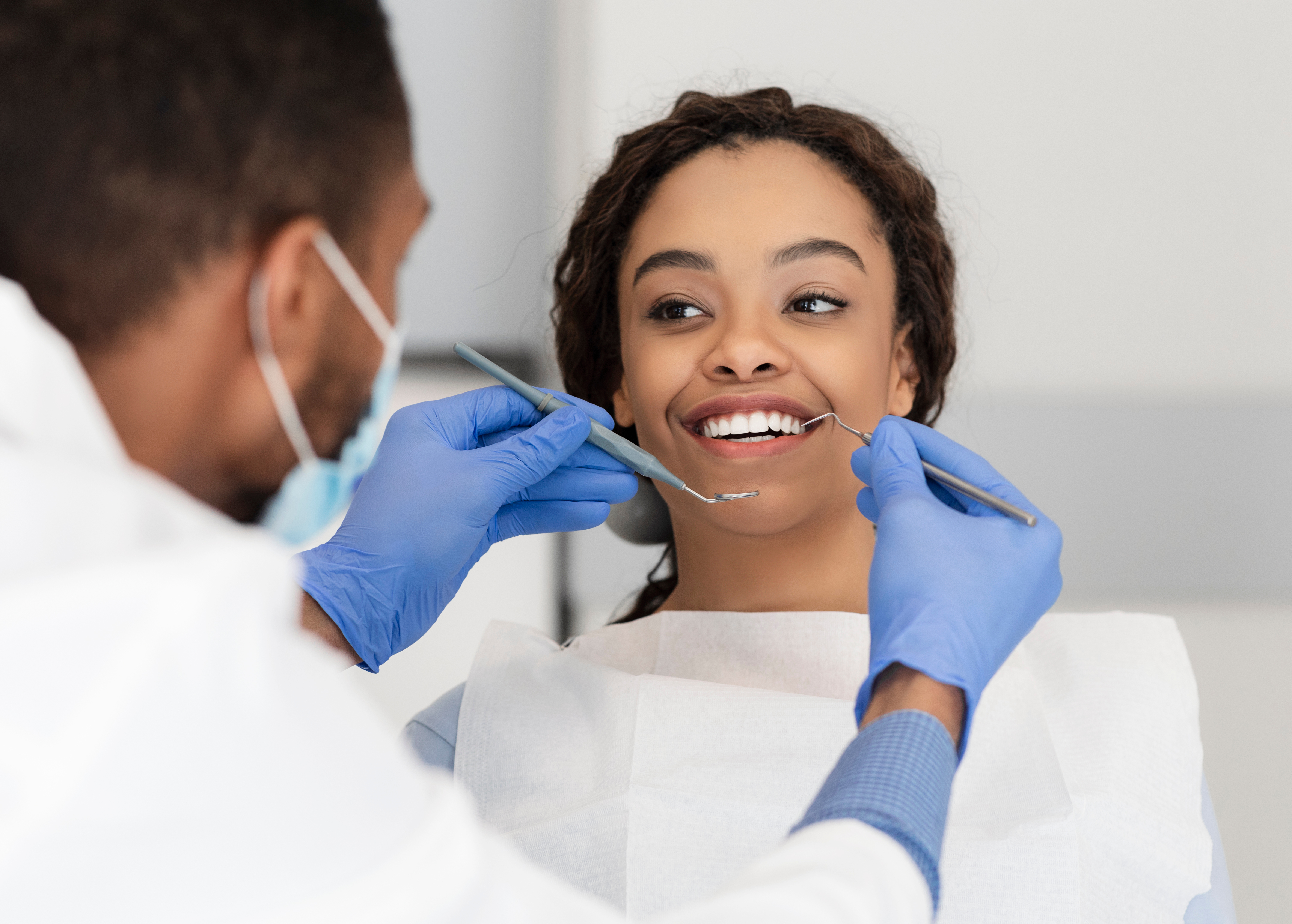 Smocked patient reclines in dental chair, smiling as masked and gloved Philadelphia prosthodontist starts to examine her teeth.