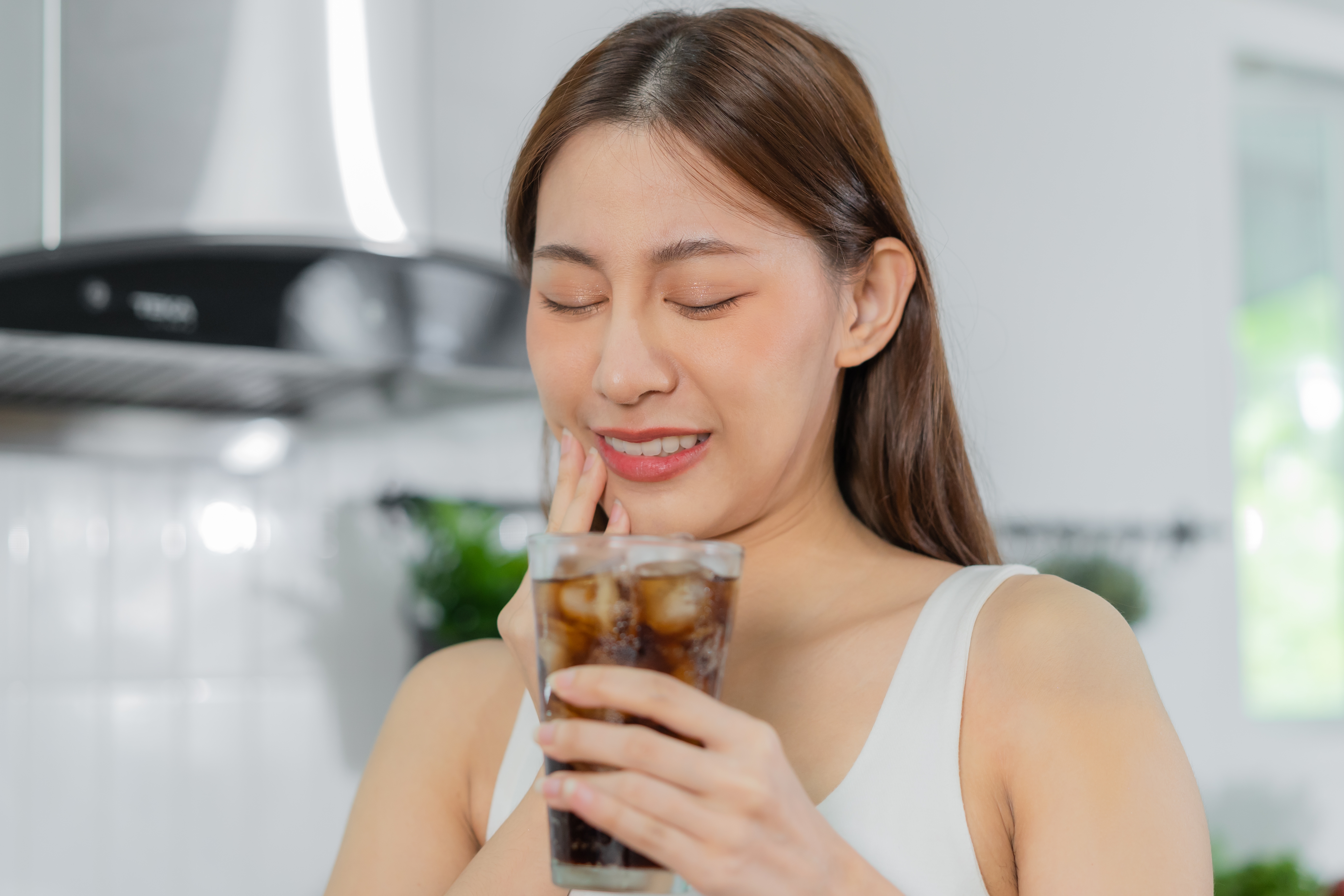 Young Asian woman holds her cheek in pain while drinking sugary soda.