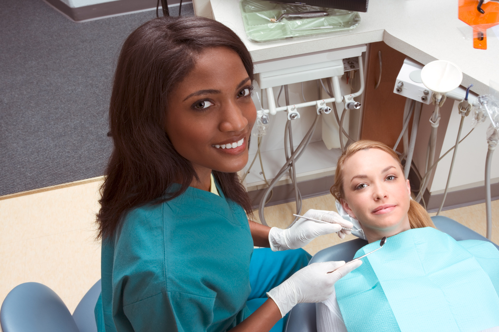 Gloved professor at dental faculty practice prepares to use a dental pick and mirror to examine the teeth of a woman in a dental chair.