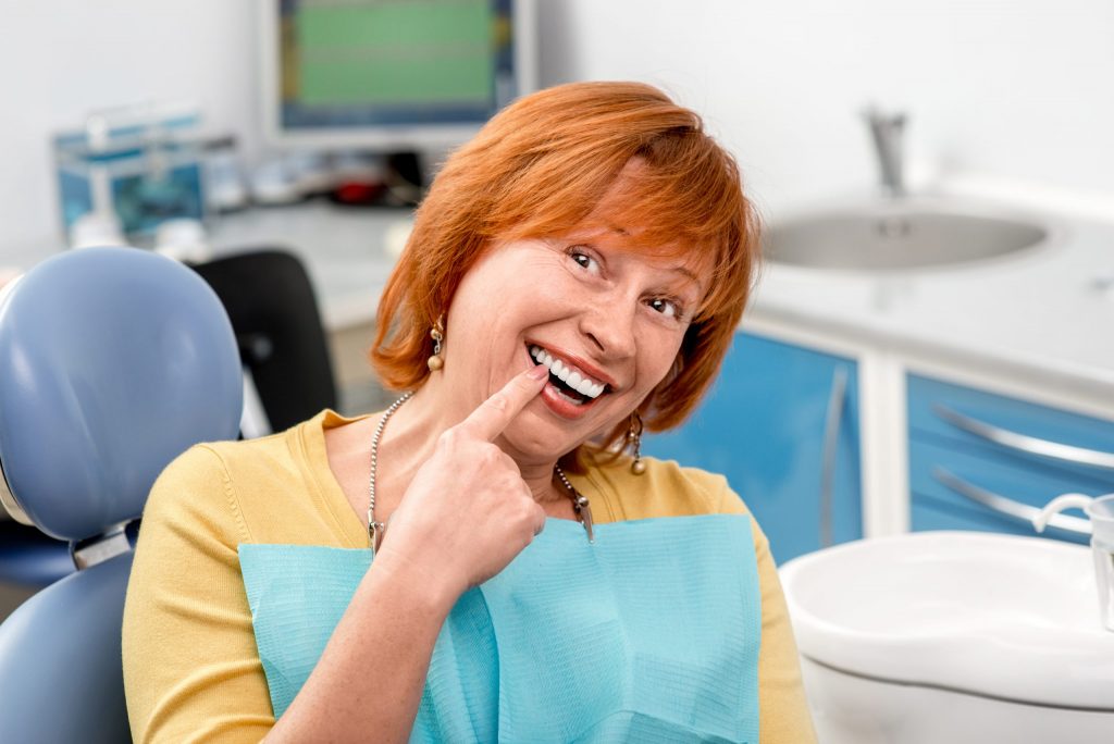 Woman sits in dental chair and smiles as she points with right index finger to chipped tooth repaired by bonding procedure.