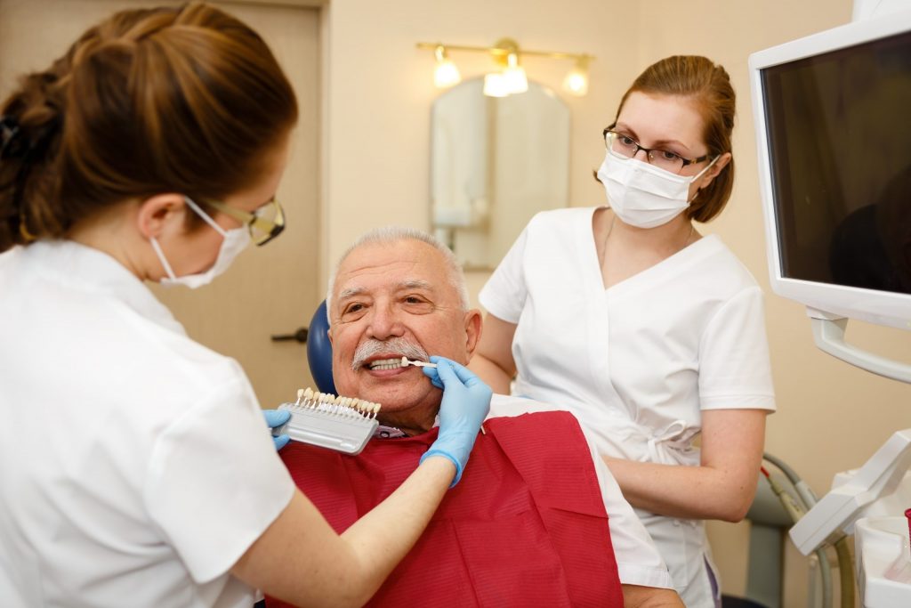 Dentist holds veneer tooth next to front tooth of man sitting in dental chair for color comparison as another dentist observes.
