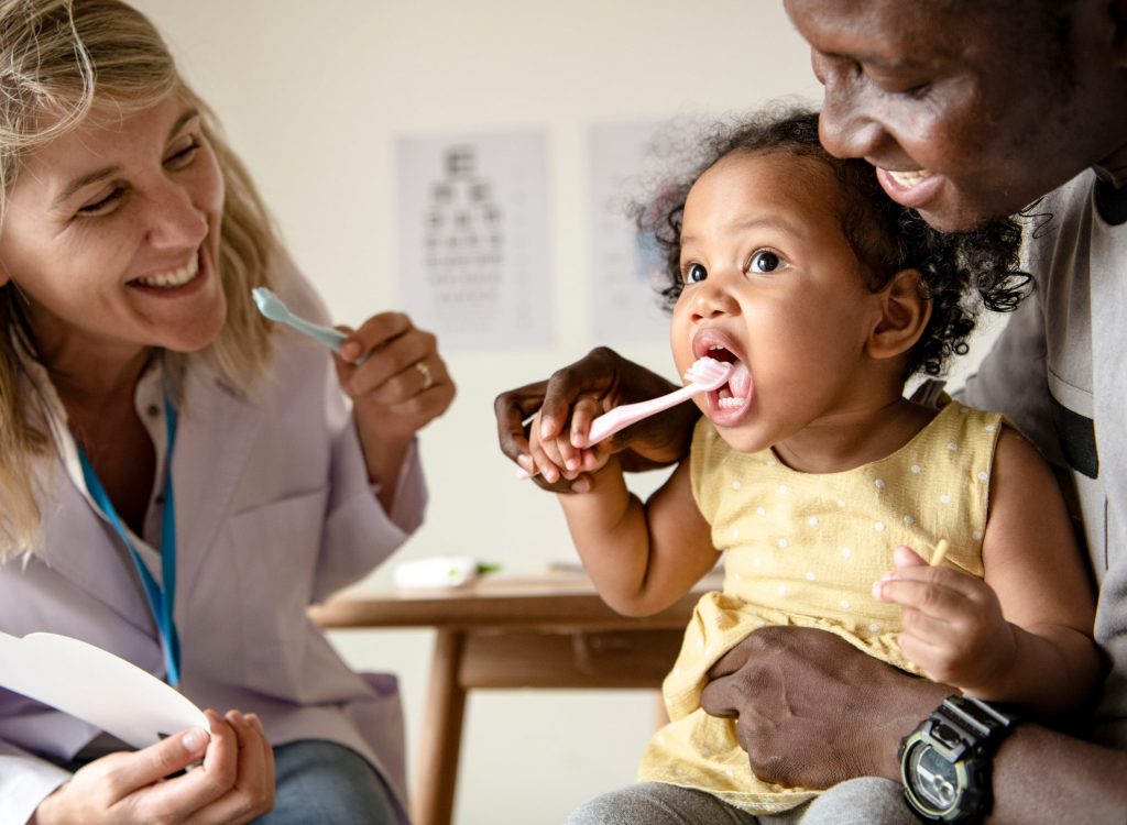 A pediatric dentist teaches a toddler how to brush her teeth.