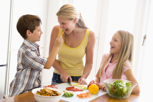 Young mother prepares mealtime accompanied by her daughter and son, all three smiling as the son offers her a carrot. 