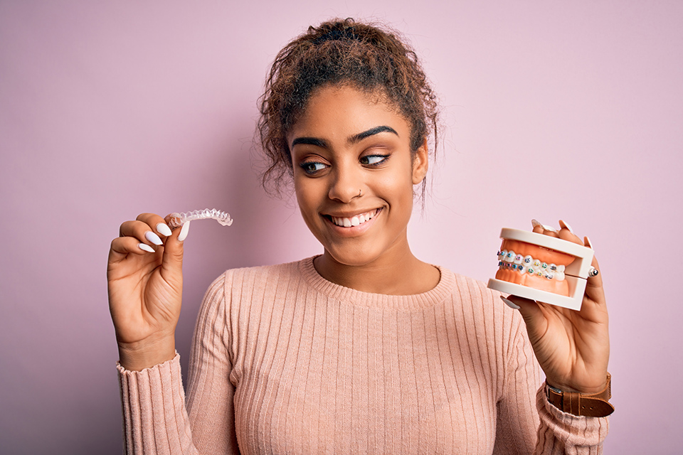 Female black teenager wearing pink shirt holding orthodontic clear aligners in one hand and mouth model with braces in other.