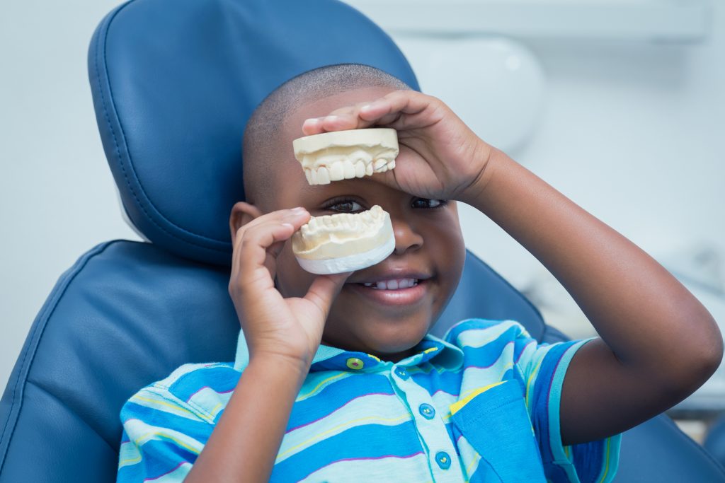 Young black male child wearing blue-striped shirt sitting in orthodontist chair holding a model of a mouth.