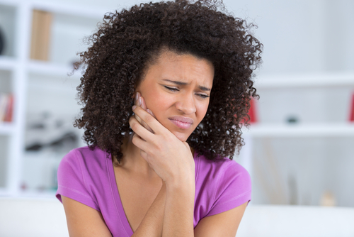 An attractive African American woman with brown curly hair, wearing a purple shirt holds her jaw in pain.