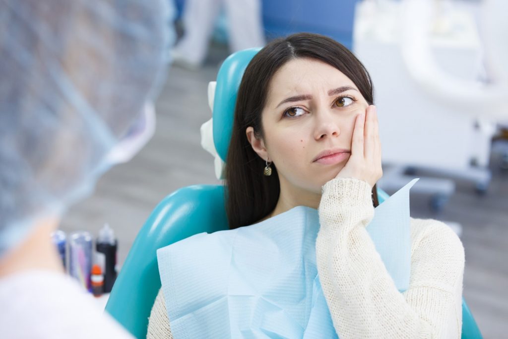 A young woman at the dentist’s office with her hand to her mouth listens to the dentist talk about depression and teeth.