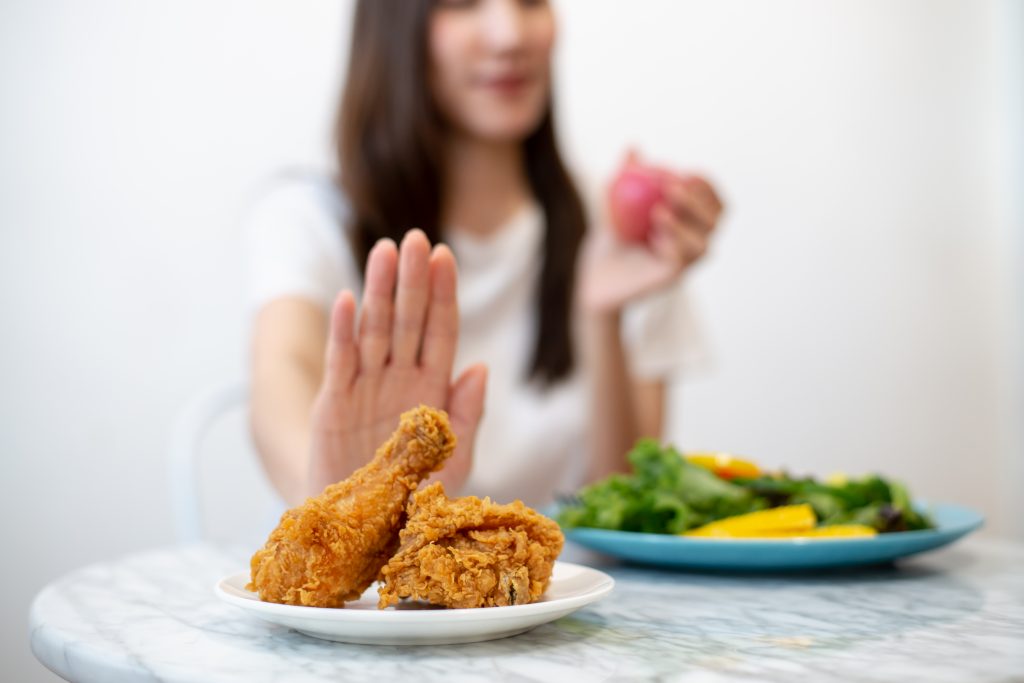 A blurred image of a woman in front of a plate of healthy vegetables and fruits shoves plate of fried chicken away.