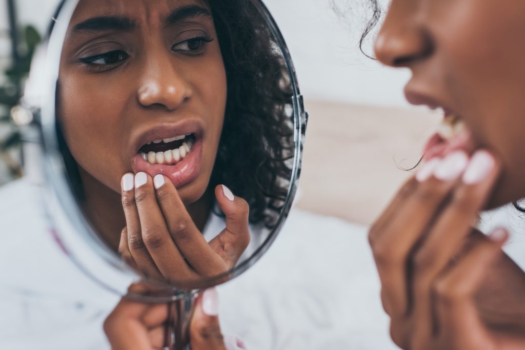 A young black woman examines her tooth enamel in a handheld mirror.