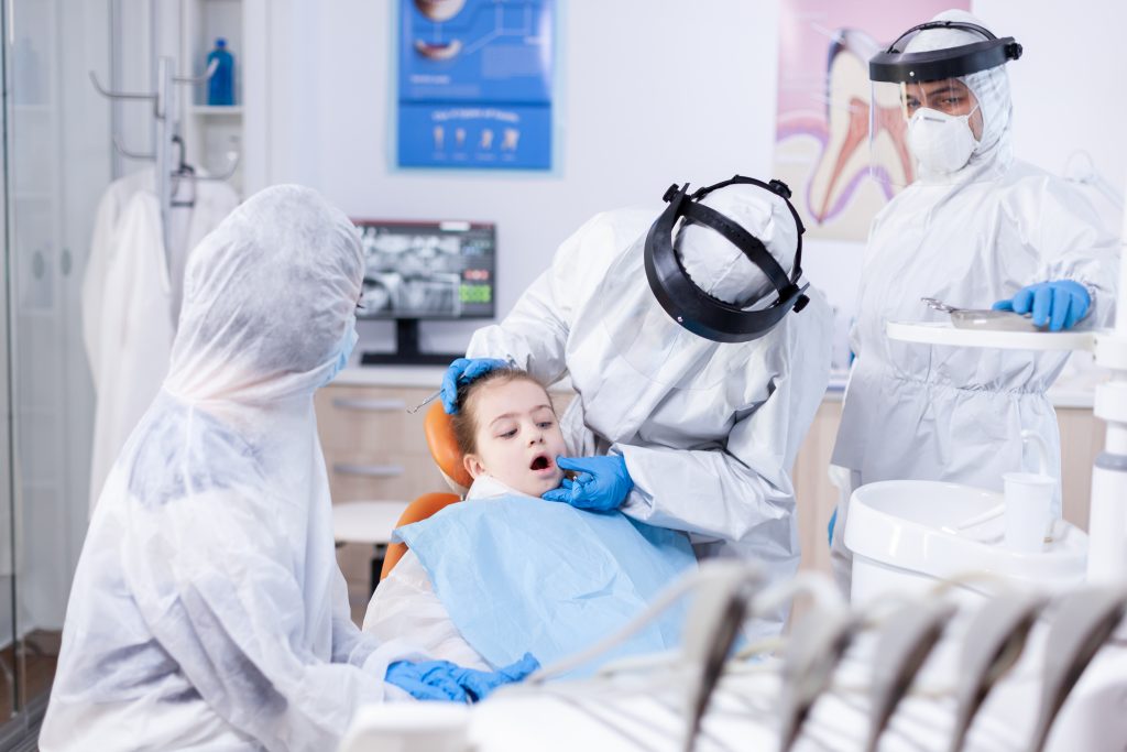 Several dentists in full personal protective equipment PPE are providing dental care to a young female patient. 