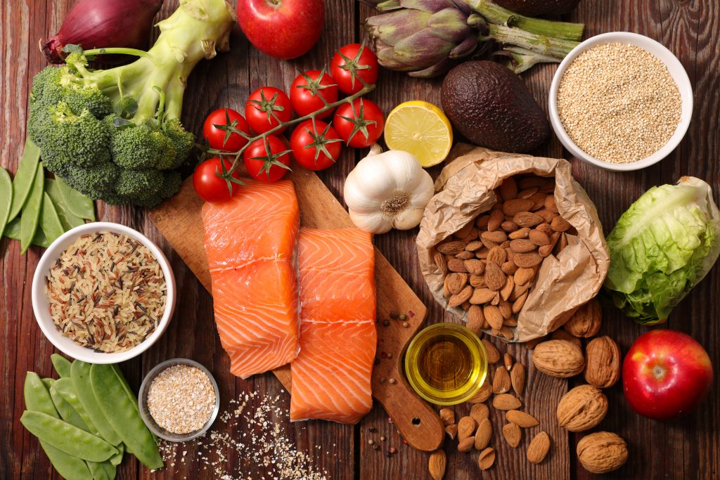 A wood table showcases healthy foods such as whole grains, fish, broccoli, nuts, peas, tomatoes, lettuce, and more.