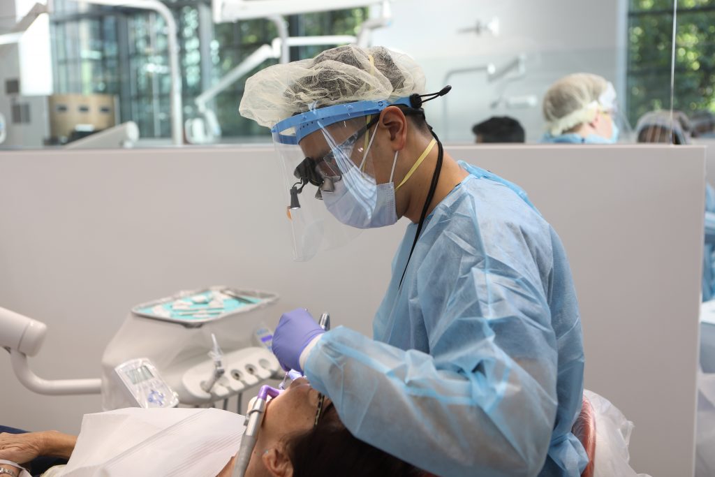 A dentist in Philadelphia wearing scrubs, face shield, and mask inspects mouth of patient reclining in dental chair.