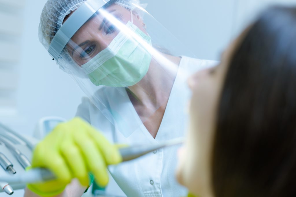 A dentist wearing scrubs, face shield, mask, and gloves shines a dental light inside patient’s mouth during dental care.