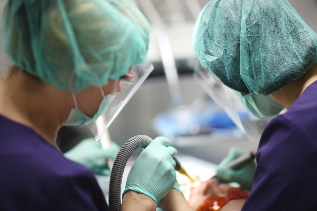 Two dentists wearing personal protective equipment (PPE) and face shields perform oral surgery on a dental patient.