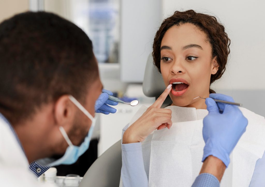  Woman sitting in dental chair points to location of a dental abscess for her masked dentist, who holds dental pick and mirror.
