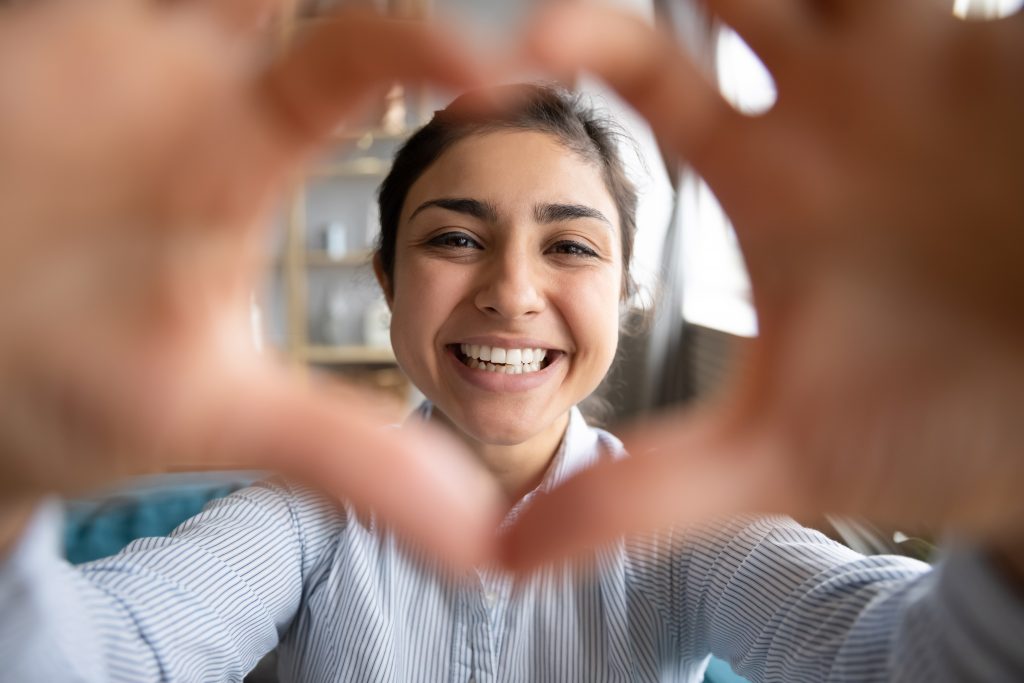 Young Indian female makes a heart out of her hand to frame her face with perfect smile from cosmetic dental care. 