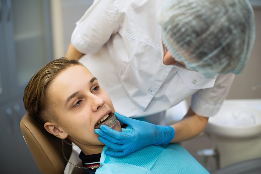 Female dentist wearing personal protective equipment (PPE) helps fit a young male patient with a mouthguard for bruxism.