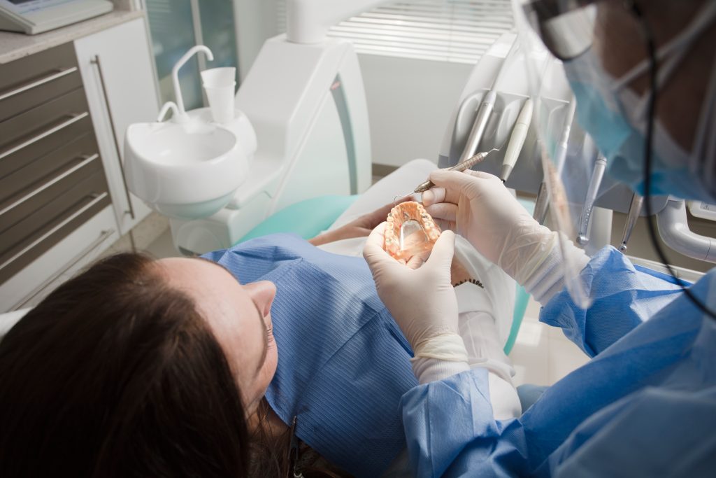 Dentist wearing personal protective equipment (PPE) explains teeth grinding symptoms to a female patient using mouth model.