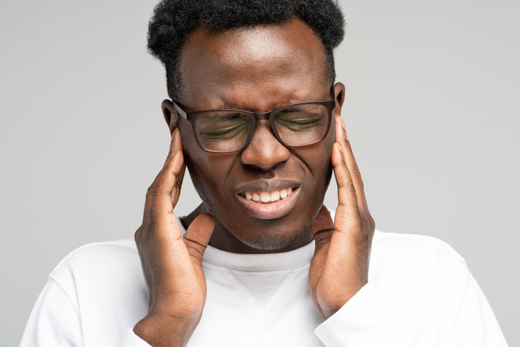 Black man wearing glasses holds his temple area from stress-related teeth grinding pain. 