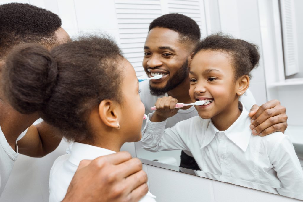 A Black father and daughter brush their teeth together.