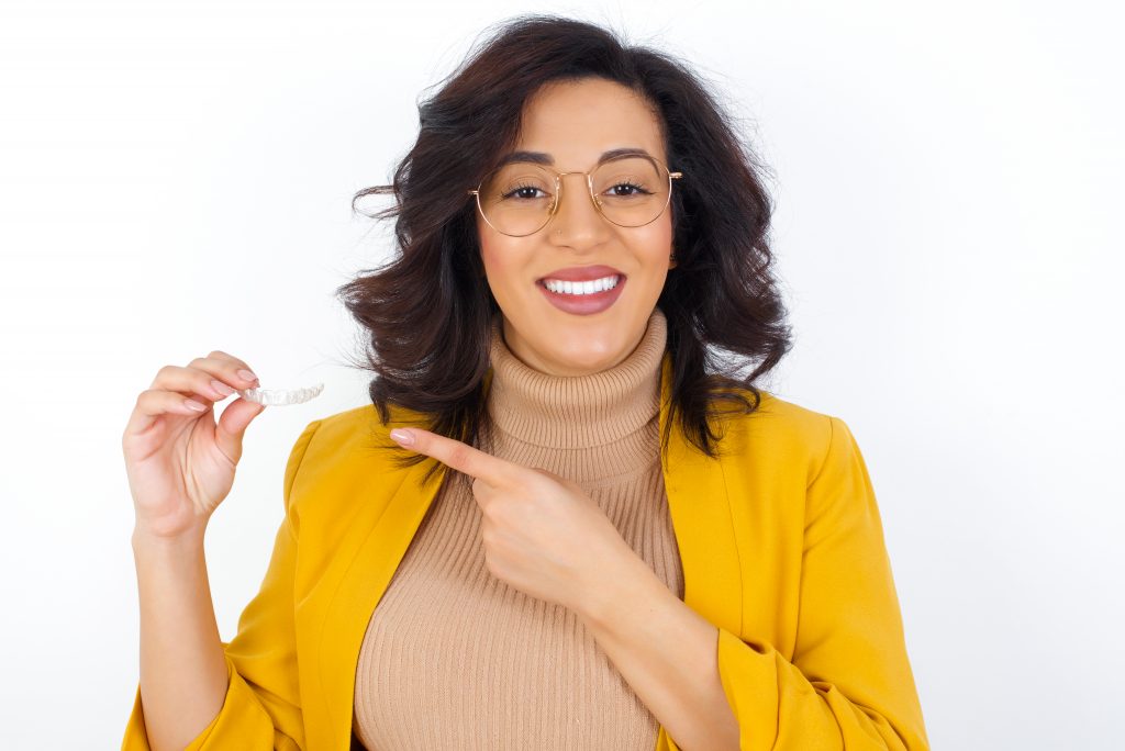 Black woman wearing glasses, tan shirt, and yellow blazer holds invisible braces using other hand to point to aligner.