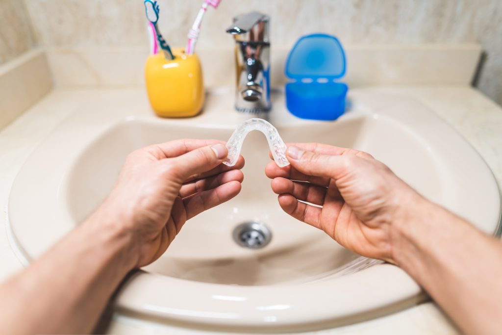  Male hands hold a clear tooth aligner over a sink getting ready to clean it. 