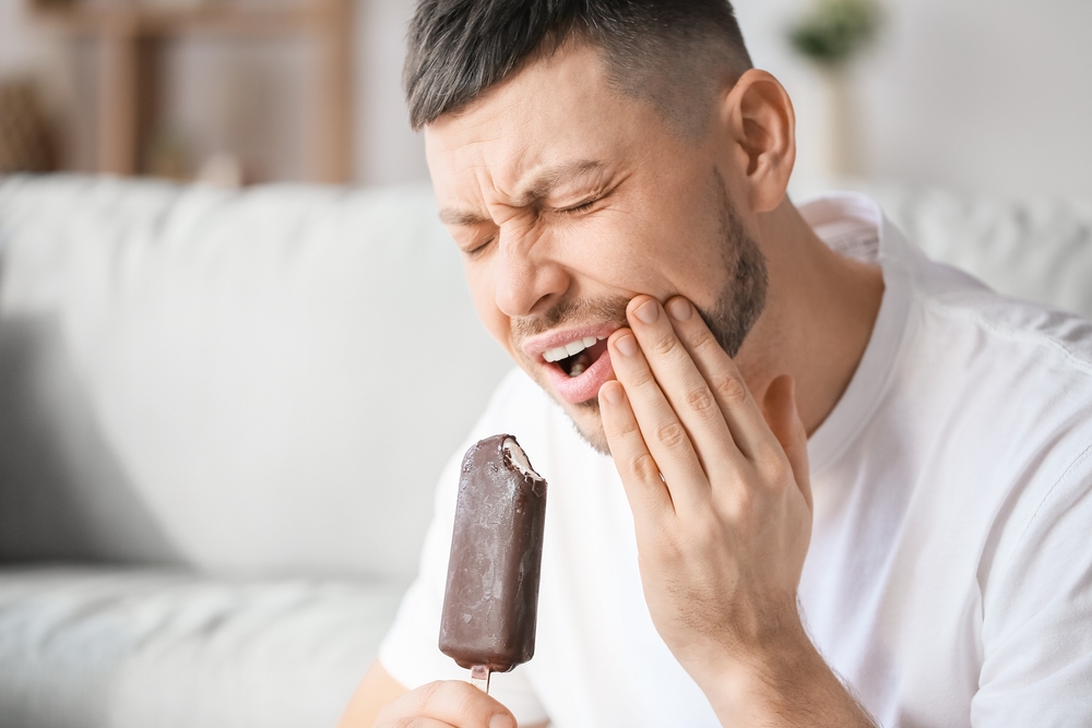 A man bites an ice cream bar and holds his hand to his mouth because he has several teeth hurting at once.