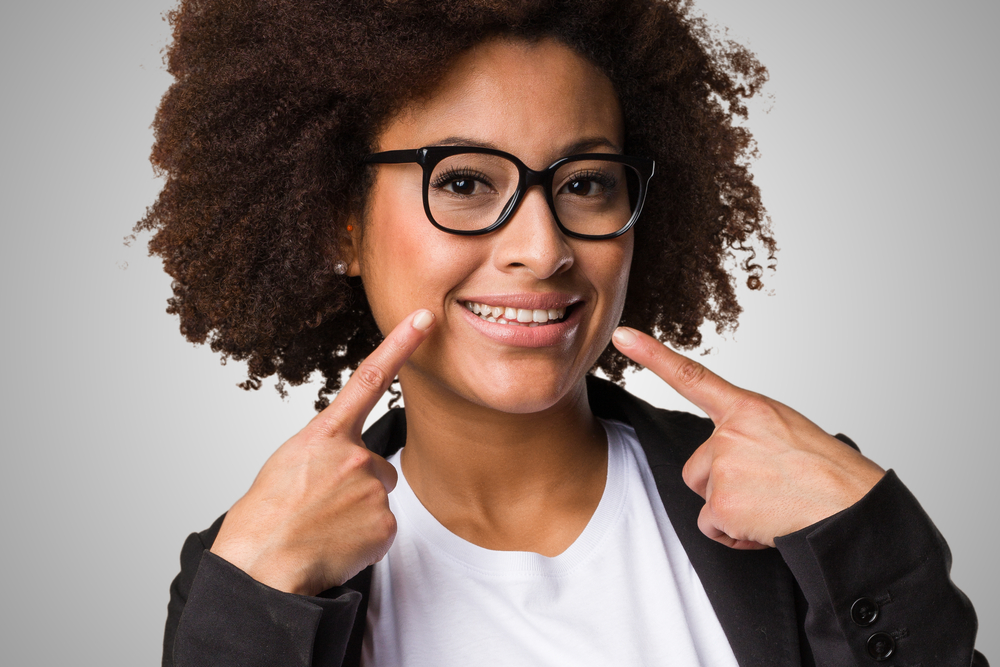  A young woman in glasses smiles and points at her teeth because she no longer has several teeth hurting at once.