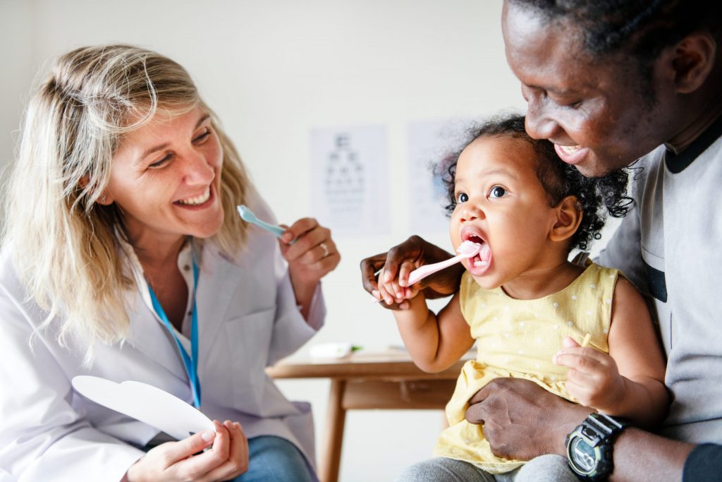 A dentist looks on while a mother helps her toddler brush her own teeth.