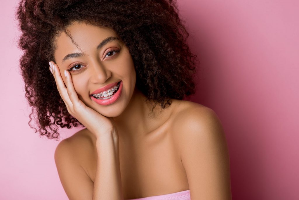 Smiling confident teen girl with metal braces poses against a pink background. 