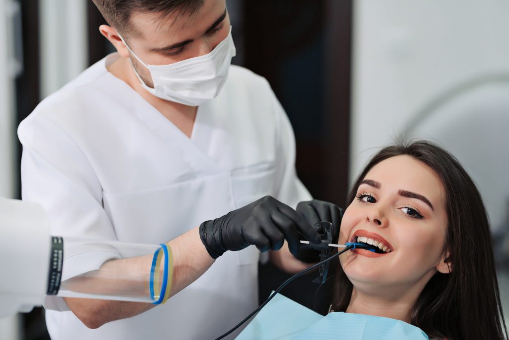 Periodontist wearing a face mask uses his dental pick to scale the teeth of a woman who reclines in a dental chair.