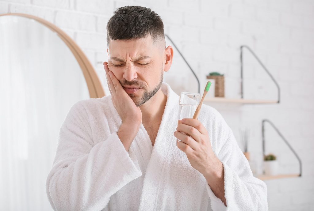  Man in bathrobe in bathroom holds toothbrush in left hand and right hand to side of jaw, feeling early stage gum disease pain.