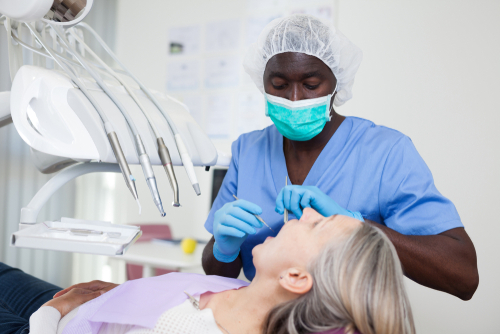 Dentist wearing hairnet, mask, scrubs, and gloves uses dental pick and mirror while inspecting mouth of woman in dental chair.