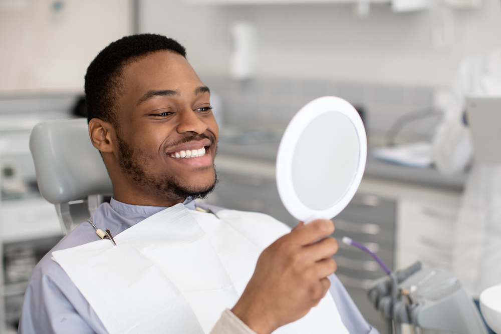 Man sits in dental chair and smiles as he looks at his reflection in handheld mirror, after dental treatment to fill cavities.