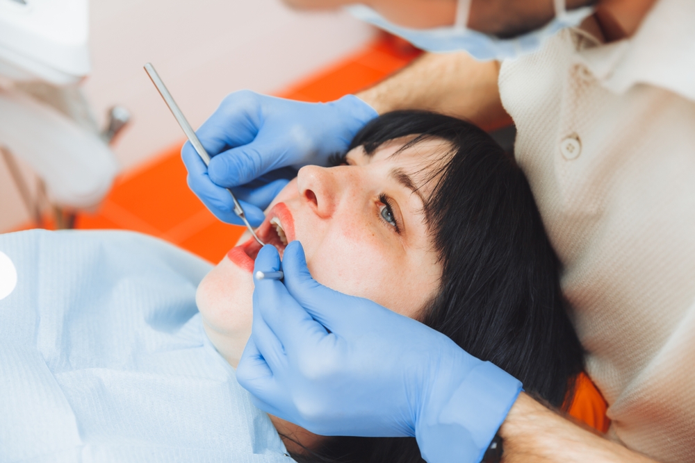 Gloved and masked dentist uses dental pick and mirror to examine mouth of woman reclining in dental chair.